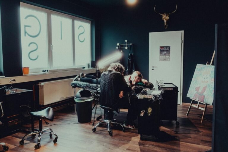 A tattoo artist while sitting on a tattoo chair is making tattoo on the back of his client lying on a tattoo bed